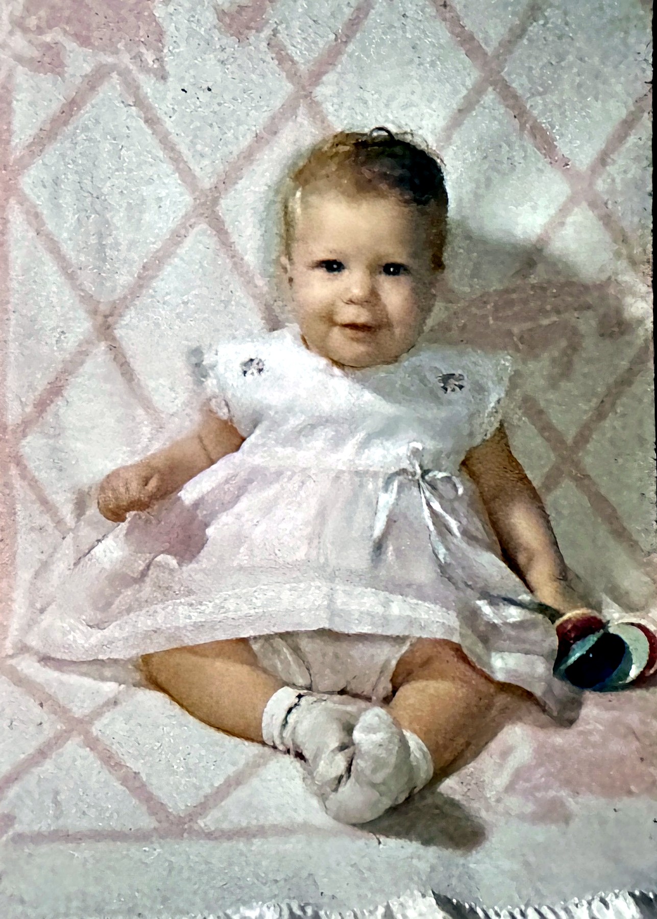 A Rubenstein baby girl in a white dress on a pink quilt with a rattle