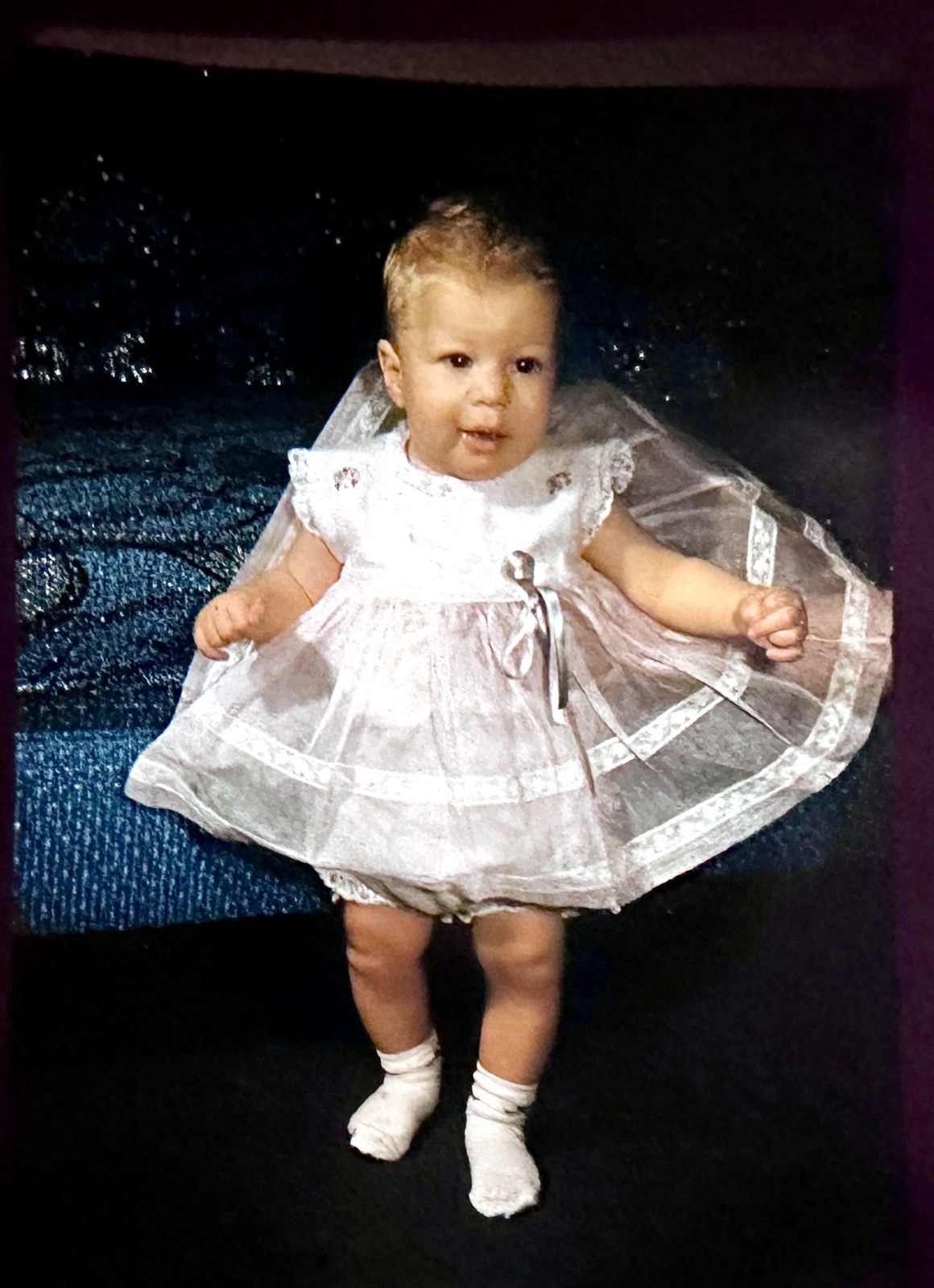 A Rubenstein baby girl standing in a lace dress on a dark blanket